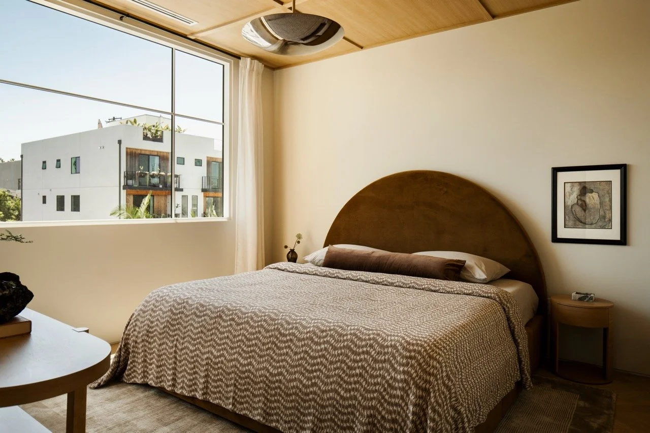 A bedroom with a large bed, wooden headboard, and beige patterned bedding next to a window with white curtains, showing an outside building with balconies.