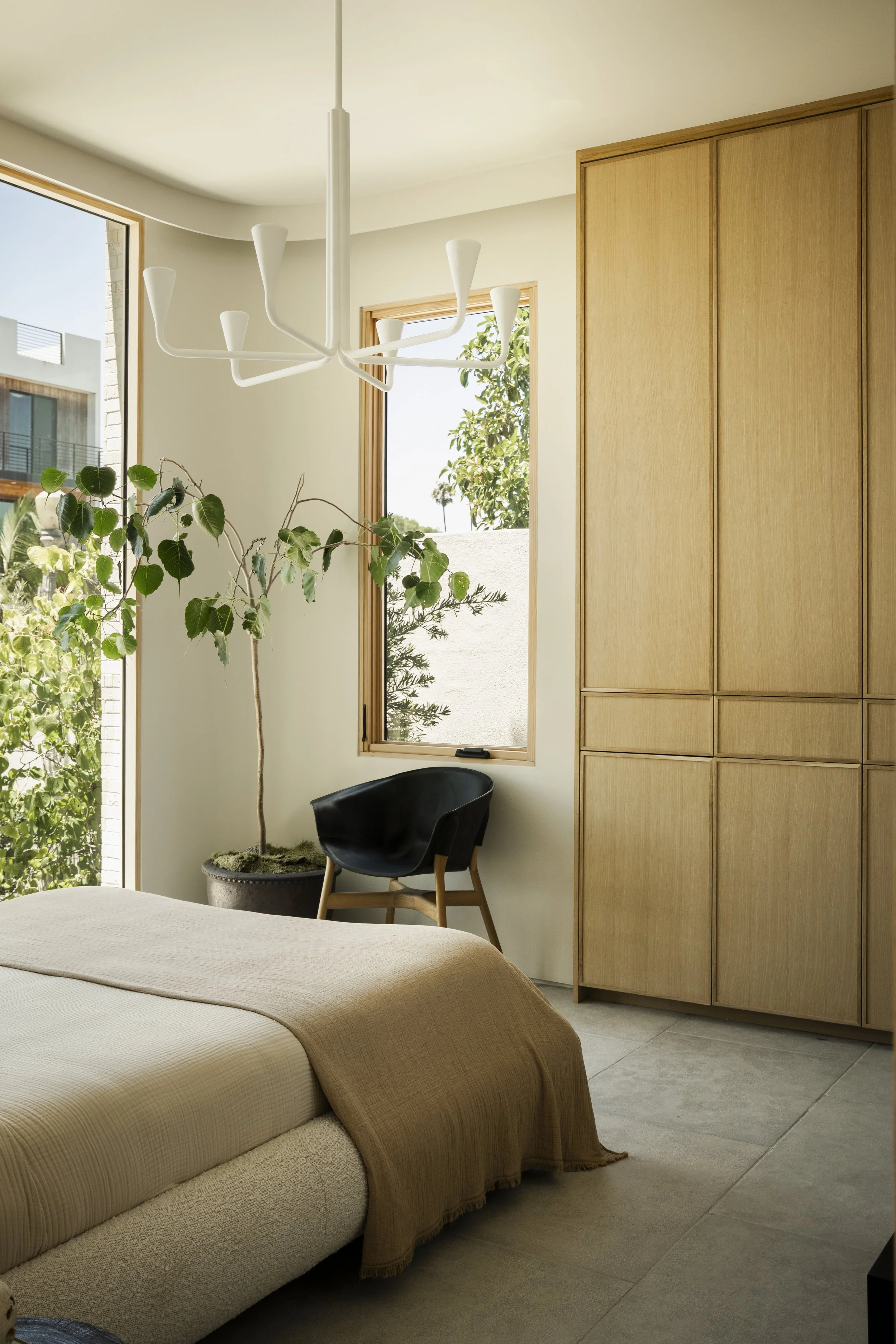 A modern bedroom with a beige bedspread, a black accent chair, and a large wooden wardrobe. There are two windows revealing trees and neighboring buildings outside. A white chandelier hangs from the ceiling, and a tall potted plant is near the window