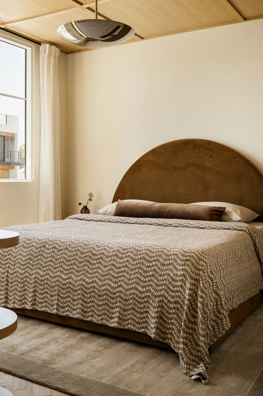A bedroom with a large bed covered in a patterned beige and brown blanket, brown and white pillows, a small side table with a vase, a window with cream curtains, and a ceiling light fixture with a reflective metallic finish.