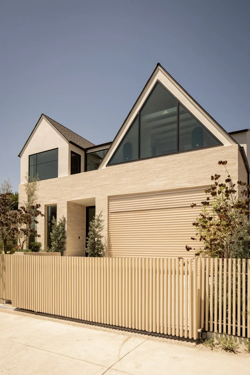 Modern two-story house with large triangular glass windows, beige brick exterior, and a beige vertical slat fence.