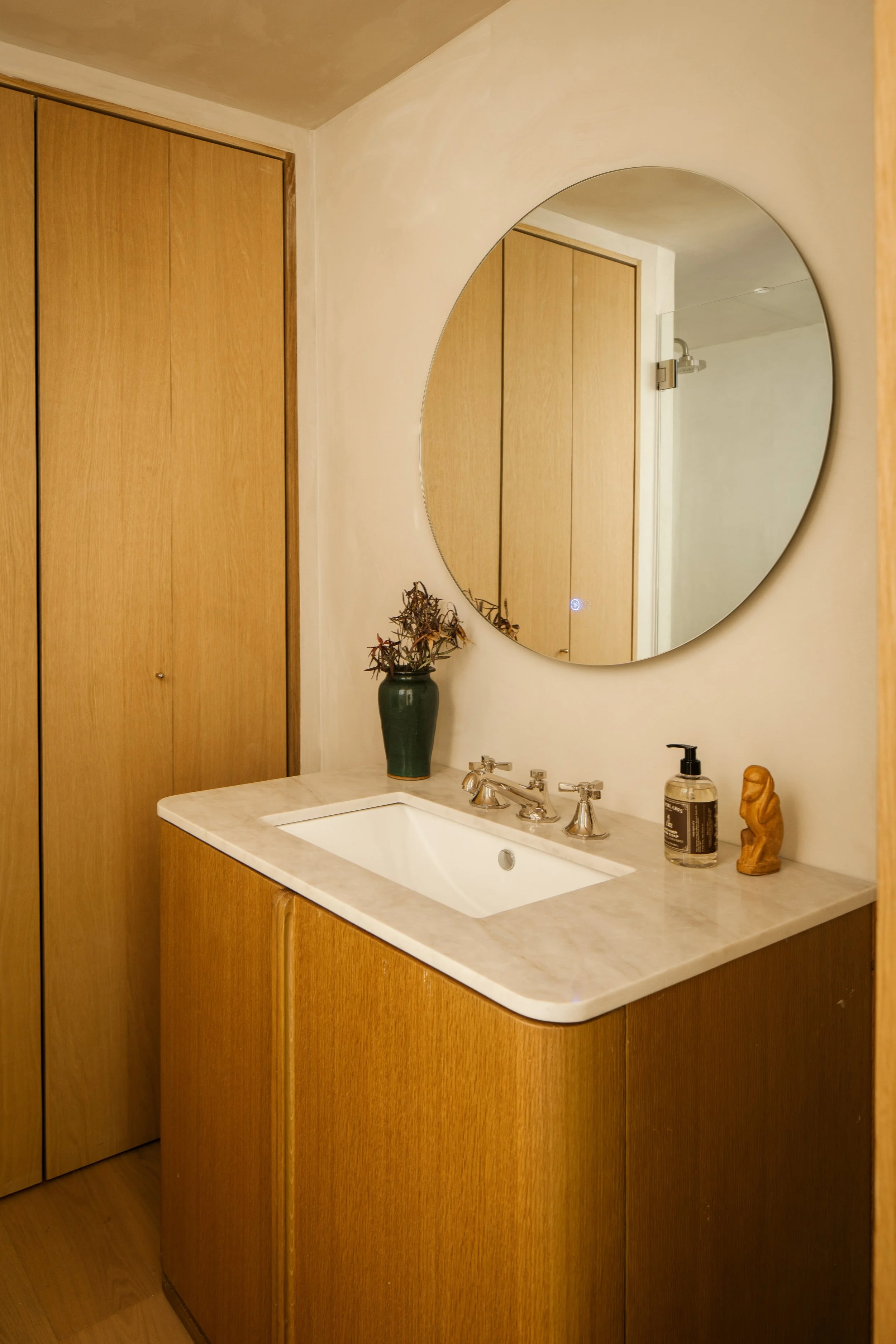Bathroom vanity with a large oval mirror, wooden cabinets, a white marble countertop, a sink, and decorative items including a green vase with dried flowers, a bottle of hand soap, and a small wooden sculpture.