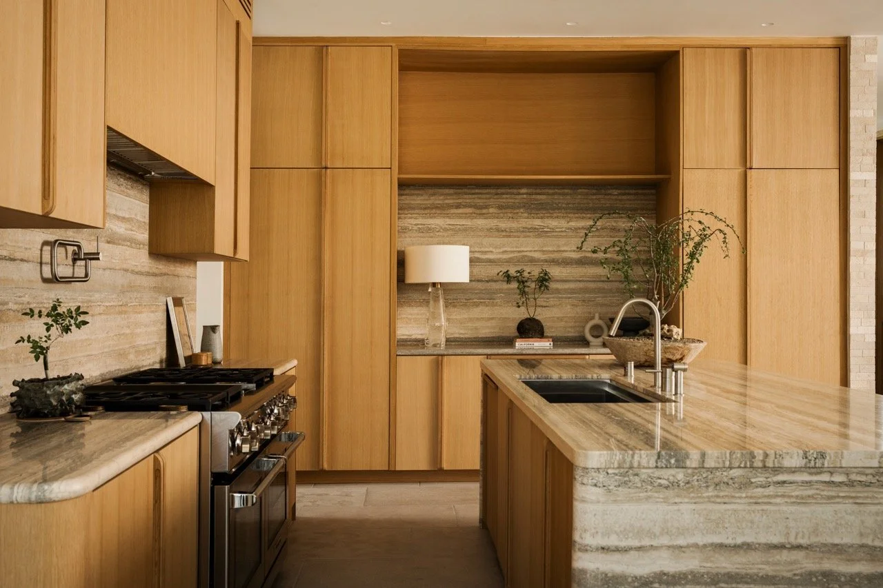 Modern kitchen with wooden cabinets, marble countertops, and a stainless steel stove. Decor includes small plants, a table lamp, and decorative objects.