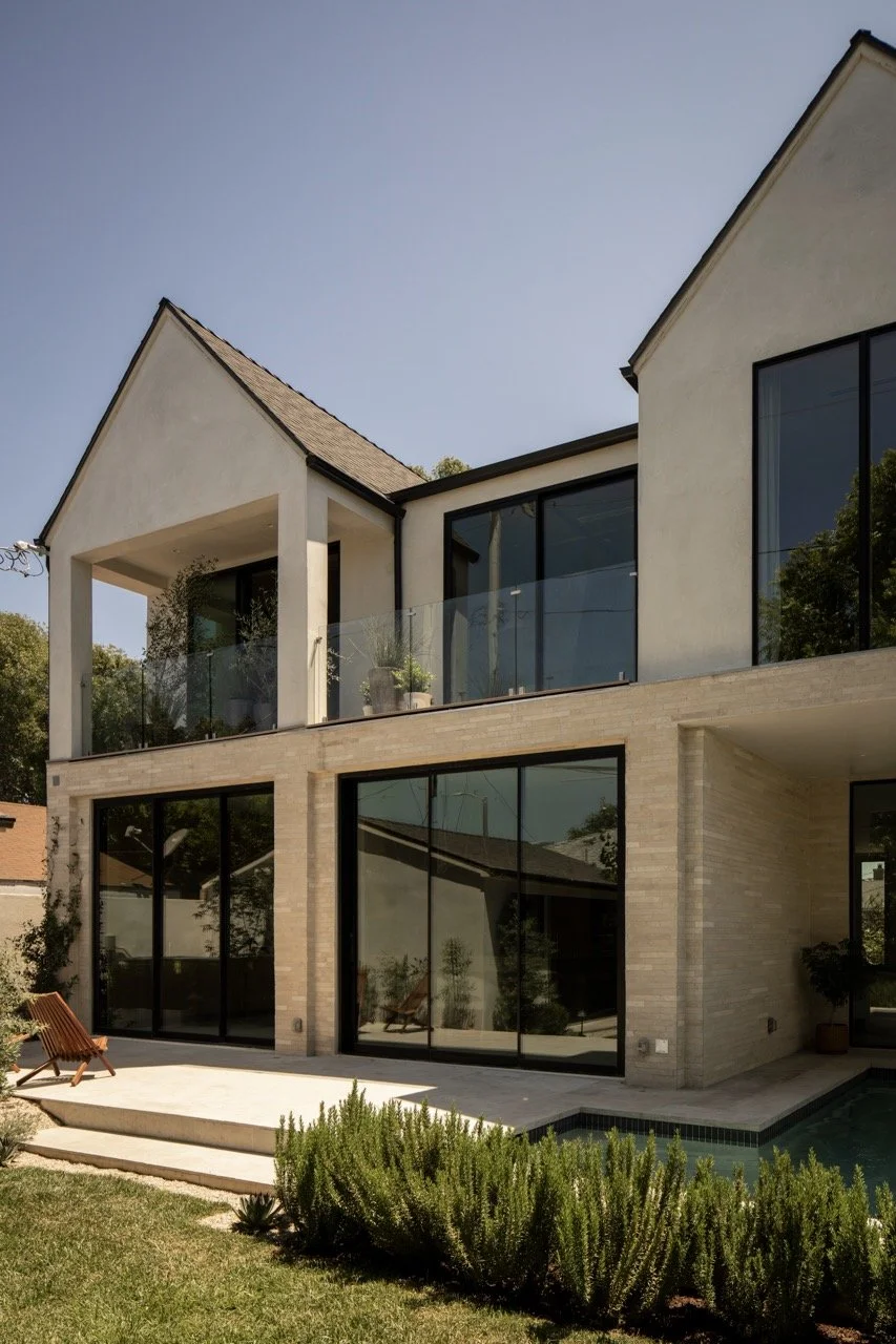 Modern multi-story house with large glass windows, a second-floor balcony with glass railing, and a patio with outdoor furniture and greenery.