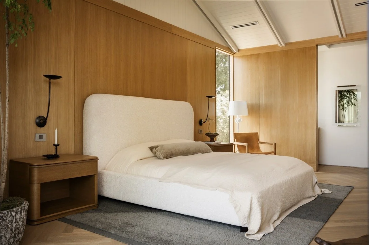 A bedroom with a white bed, beige bedding, and a beige pillow. The headboard is padded and off-white. There are wooden nightstands on both sides of the bed, each with a black wall-mounted lamp. A candle is on the left nightstand. There is a wooden ch