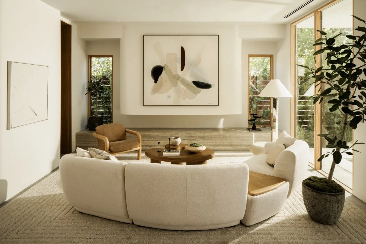 Modern living room with white curved sofa, brown armchair, wooden coffee table, large abstract art on the wall, tall window with wooden frame, potted plant, and floor lamp.
