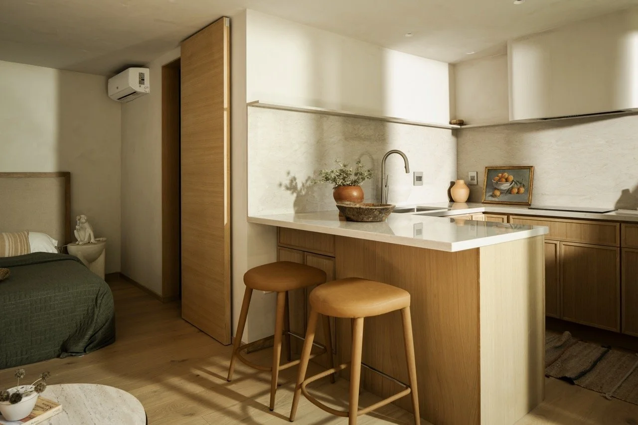 A cozy apartment interior featuring a small kitchen with a white countertop, two wooden bar stools, and decorative vases. The living area has a bed with a beige headboard, and a side table with a sculpture. There is an air conditioning unit on the wa