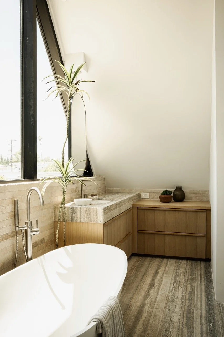 Modern bathroom with a freestanding bathtub, wooden cabinets, a marble countertop, potted plants, and large windows