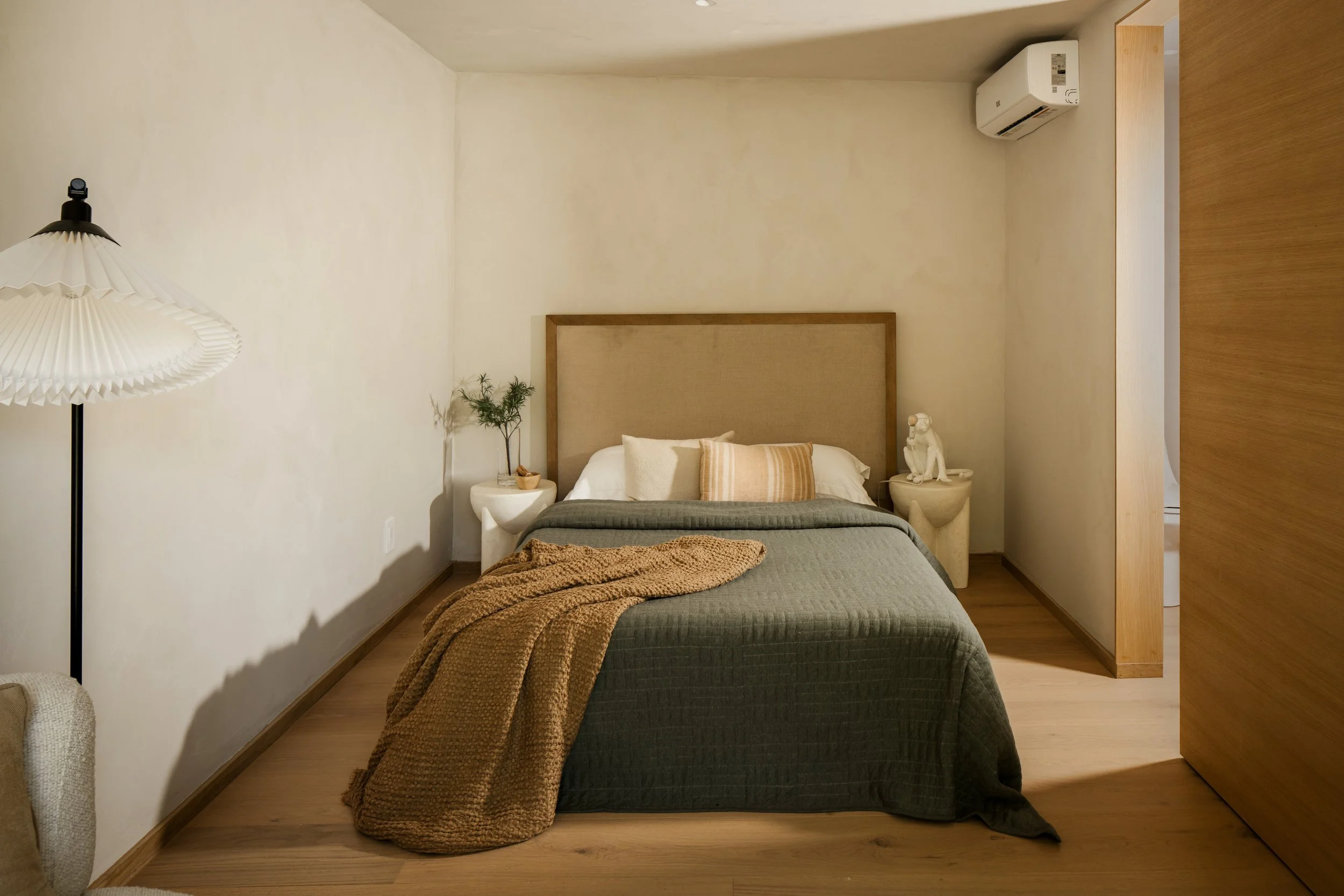 A cozy bedroom with a large bed featuring a beige headboard, white pillows, and a textured brown blanket. Beside the bed are two white side tables, one with a small plant, and the other with a decorative white dog sculpture. A floor lamp with a pleat