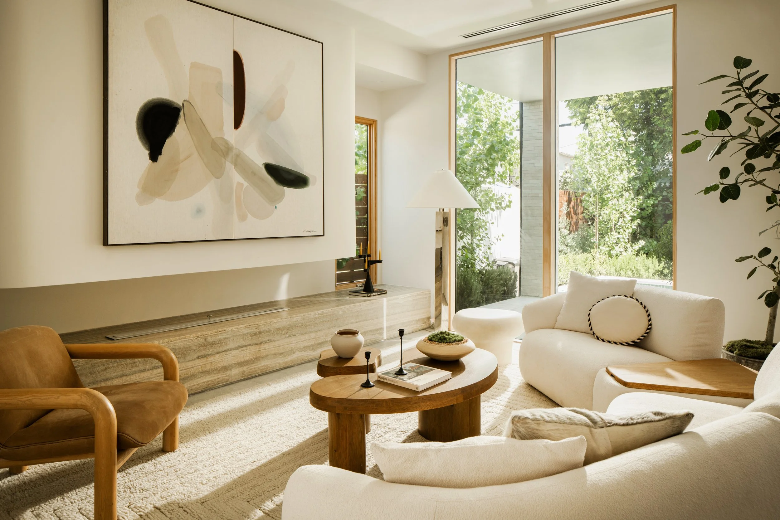 A living room with large windows, a white sofa, a round wooden coffee table, abstract artwork on the wall, a table lamp, and green plants, overlooking a garden outside.