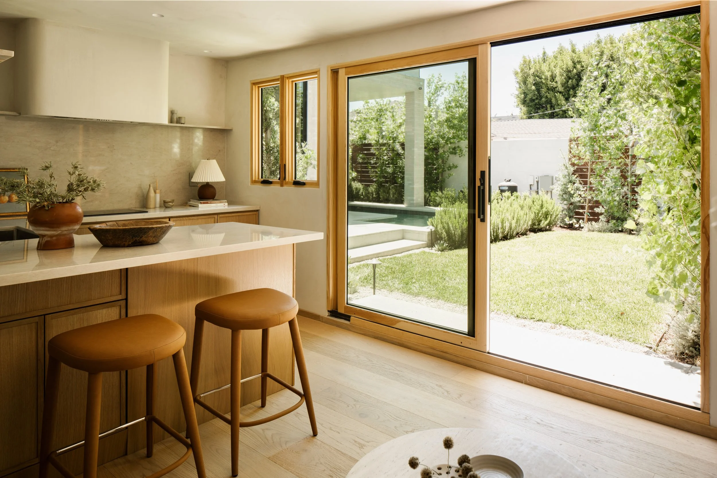The image shows a modern kitchen with a white countertop, two brown stools, and a sliding glass door leading to a lush backyard with green grass and trees.