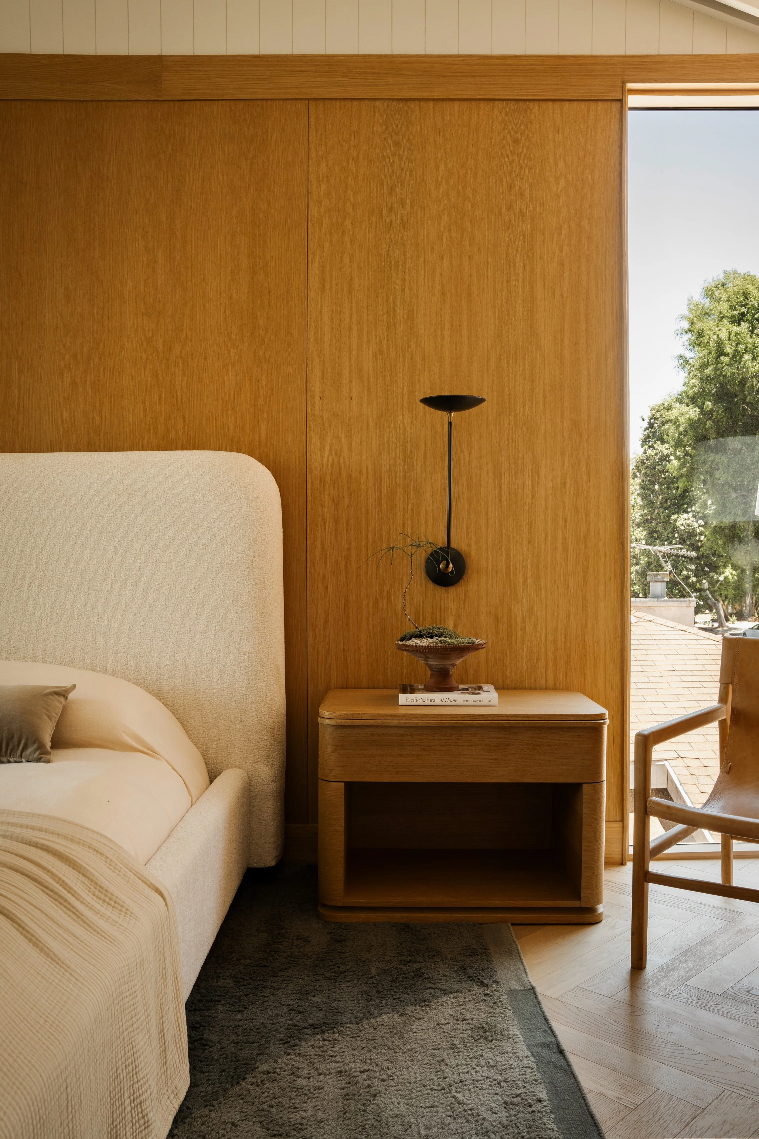 Interior bedroom with a beige upholstered bed, wooden nightstand with a bonsai plant and a book, wooden wall paneling, floor lamp, large window with view of trees and sky, and wooden chair.