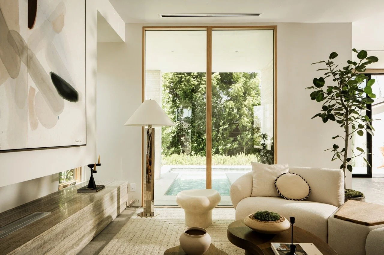 Modern living room with white sofa, large plant, glass sliding doors with views of greenery and pool outside, art piece on wall, and neutral-toned furniture.