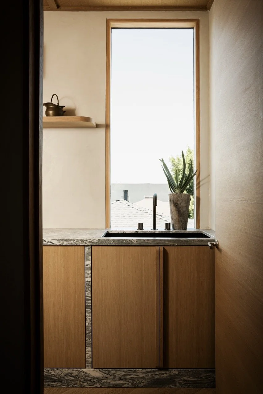 A small kitchen nook with a window, a potted plant on the sink, and a shelf with a teapot. The cabinetry is made of wood, and the countertop is a marble pattern.