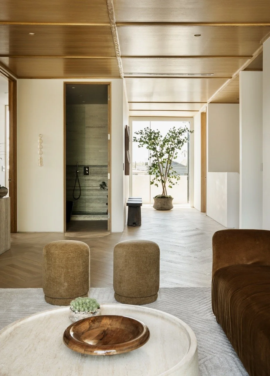 A modern living room with a wooden ceiling, beige walls, and a large window with natural light. There are two brown ottomans, a sofa, a round coffee table with a wooden bowl, a small plant, and a large potted plant near the window.