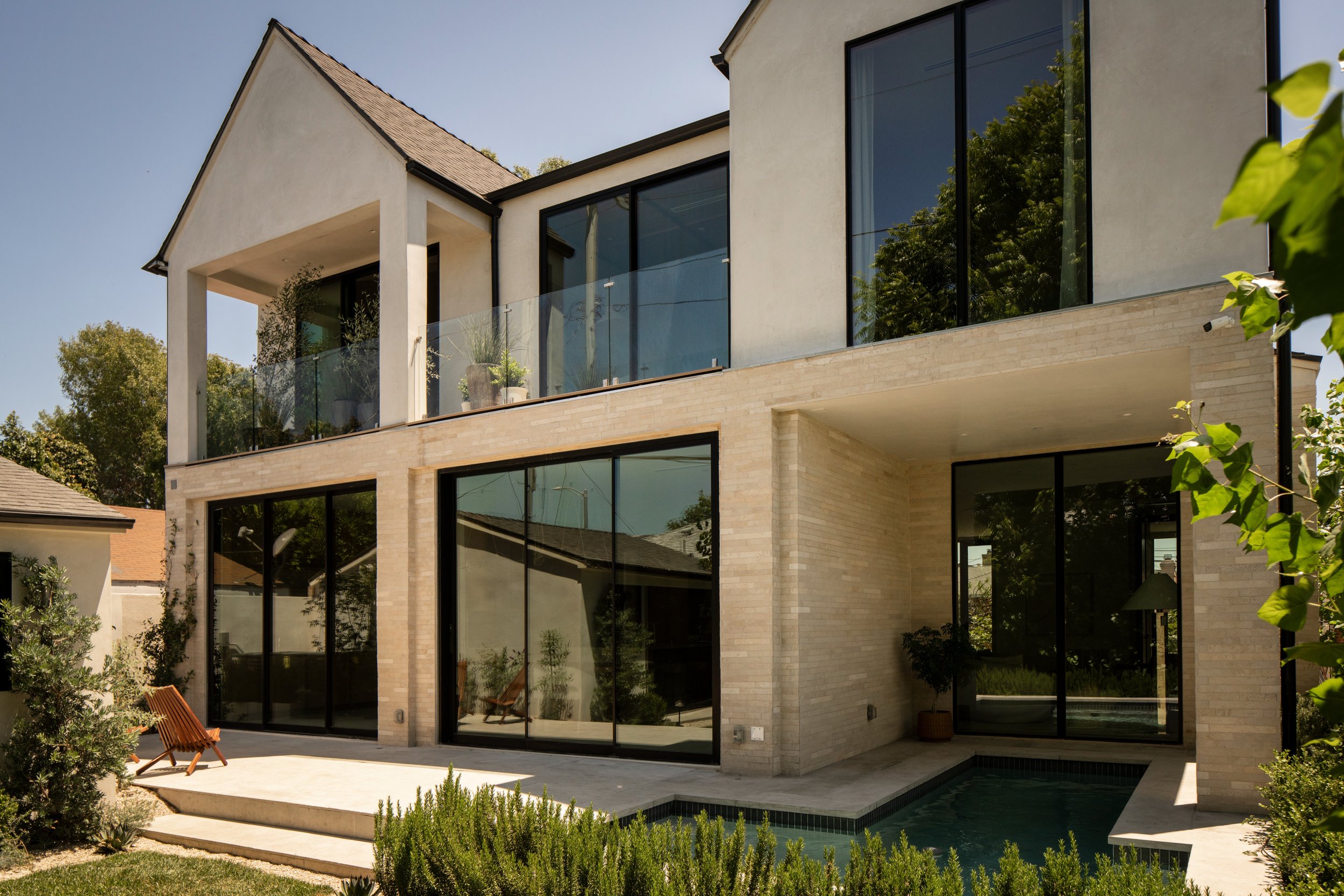 Modern multi-story house with large glass windows and sliding glass doors. The house has a beige brick exterior and a glass balcony with a plant pot. There is a small patio area with outdoor furniture and a garden with shrubs and trees surrounding th