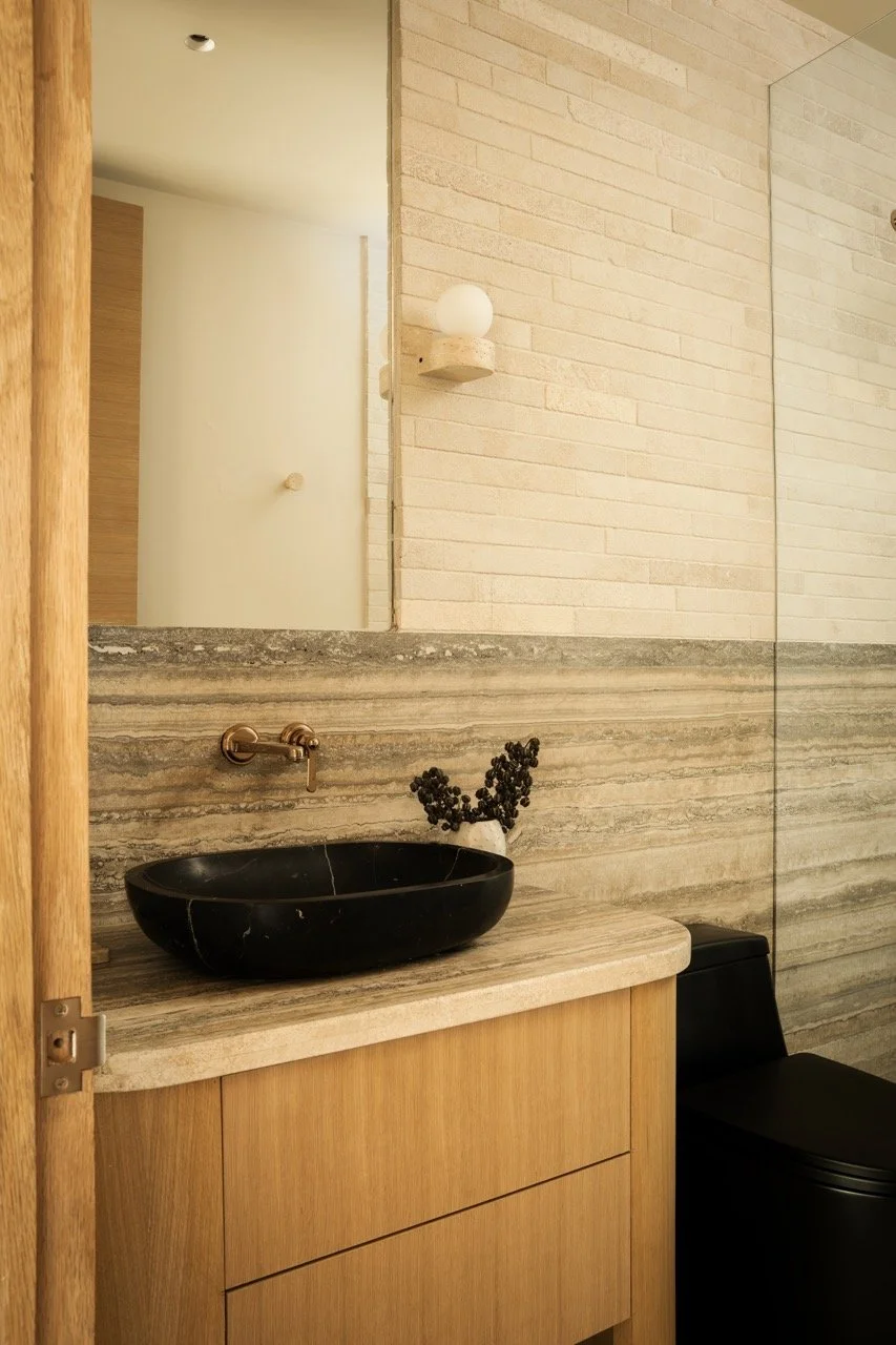 Bathroom with a black oval sink on a beige marble countertop, wooden cabinet, beige and gray striped textured wall, mirror, wall-mounted gold faucet, black toilet, and a decorative plant.