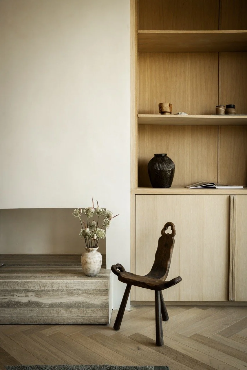 A minimalist interior with a wooden shelf filled with small decorative items, a ceramic vase with flowers on a stone surface, and a wooden chair with a carved backrest.