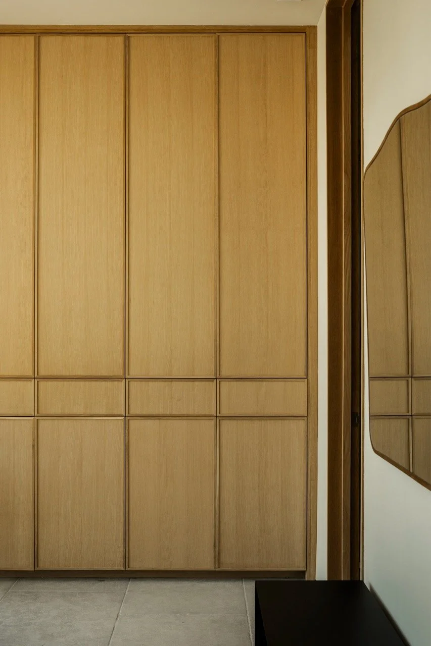 A wooden closet with multiple doors, beside a wall with a mirror and a black table or bench on a tiled floor.