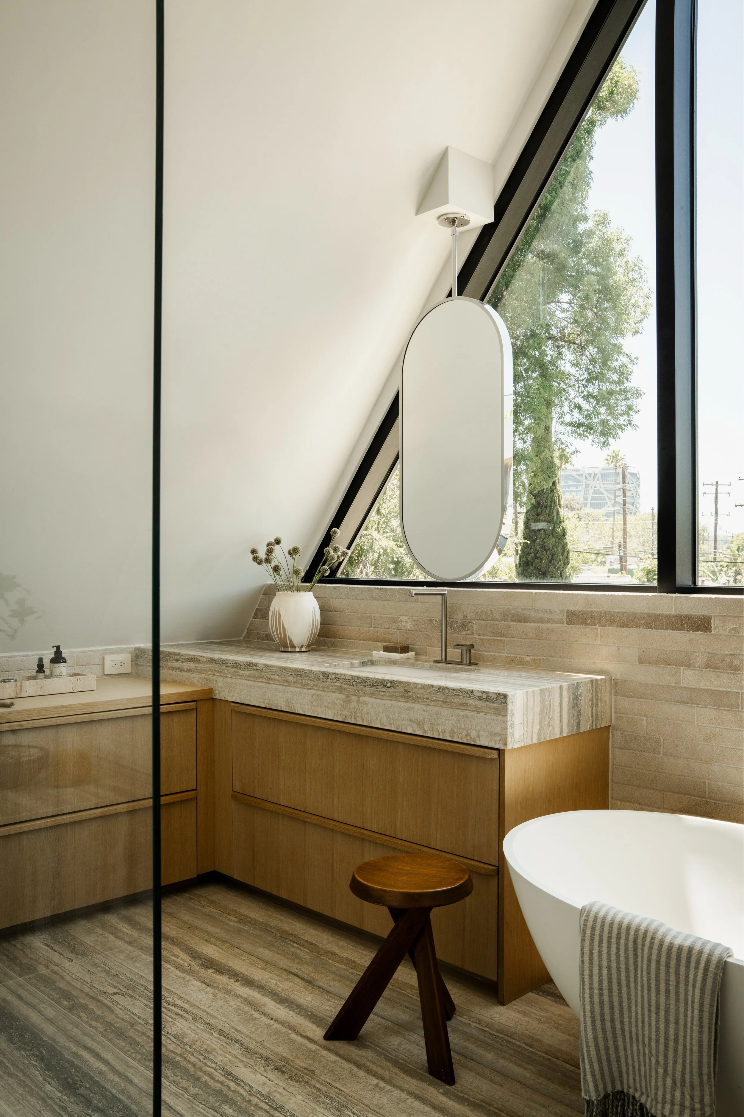 A modern bathroom with a sloped ceiling, large window with a view of trees, a mirror, a vase with dried flowers, and a bathtub with a towel draped over it. Wood cabinetry and stone countertop. Wood stool.