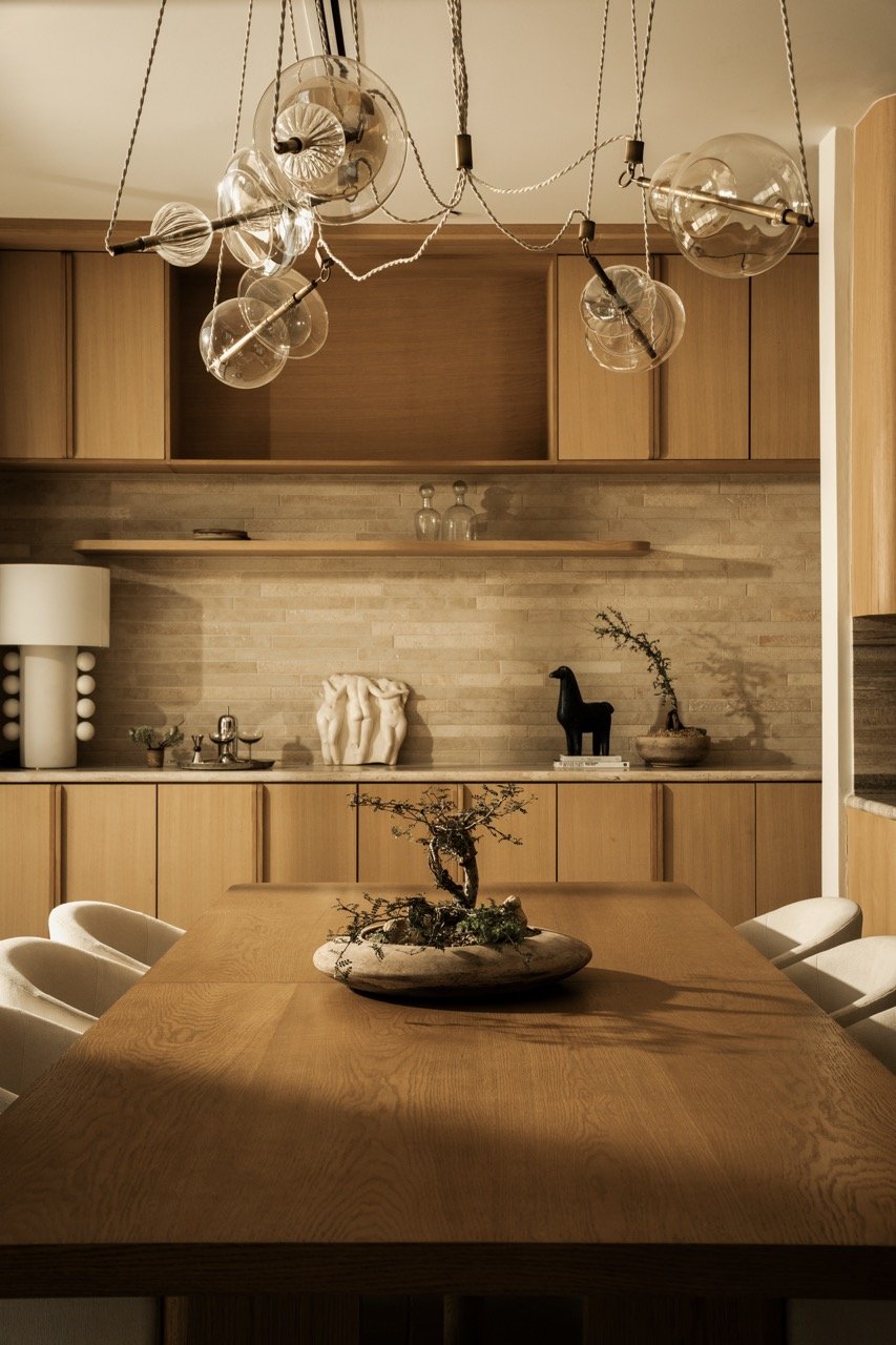 A modern kitchen with wooden cabinets, a wooden dining table with a bonsai plant centerpiece, and a decorative light fixture hanging from the ceiling.
