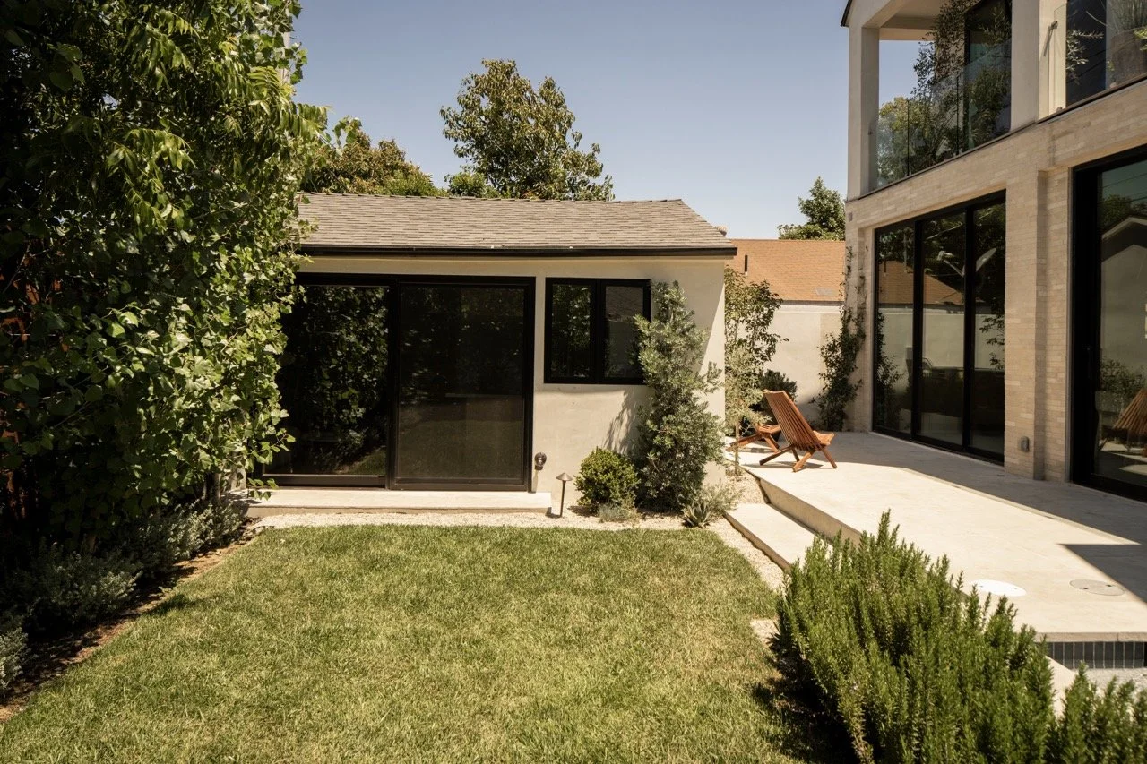View of a backyard with green grass, various plants, and trees. There is a small white building with black-framed sliding glass doors and windows, next to a larger modern house with large glass windows and a light-colored brick exterior. Wooden chair