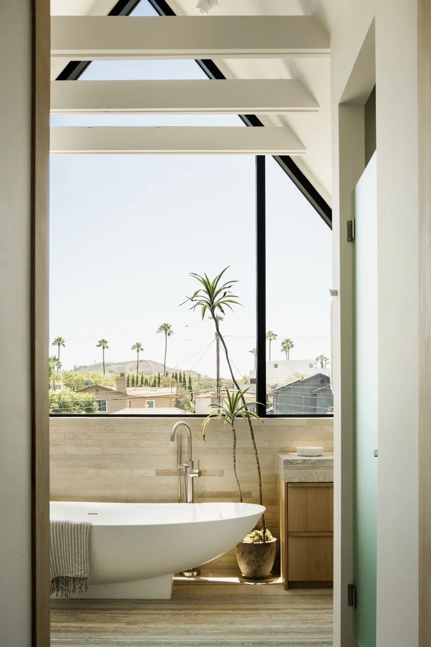Modern bathroom with a freestanding bathtub, potted plant, and large window showing a view of palm trees and a hilly landscape.