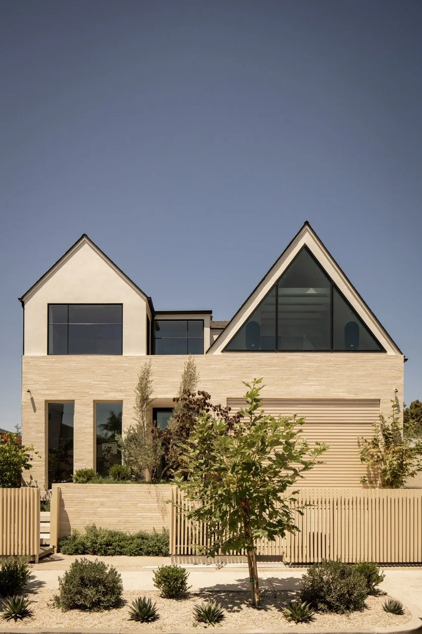 Modern house with angular rooflines, large glass windows, surrounded by landscaped garden with small trees and shrubs, beige walls, and a wooden fence, under a clear blue sky.