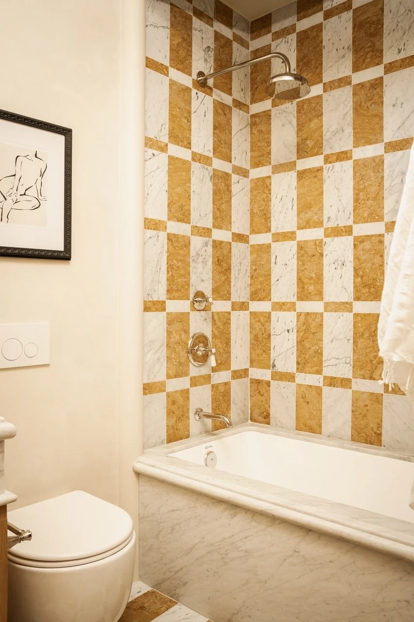 Bathroom with beige walls, framed artwork of a nude figure, and a bathtub with a square marble surround. There is a showerhead and controls on a tiled wall with a checkered pattern of beige and white tiles.