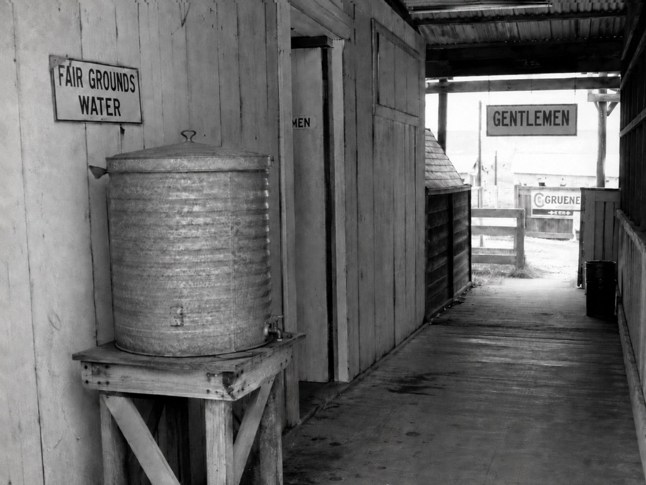 Black-and-white 1940s photograph of a Fair Grounds Park walkway with a galvanized water cooler, restroom signage, wooden walls, and a shaded passage near the grandstand.