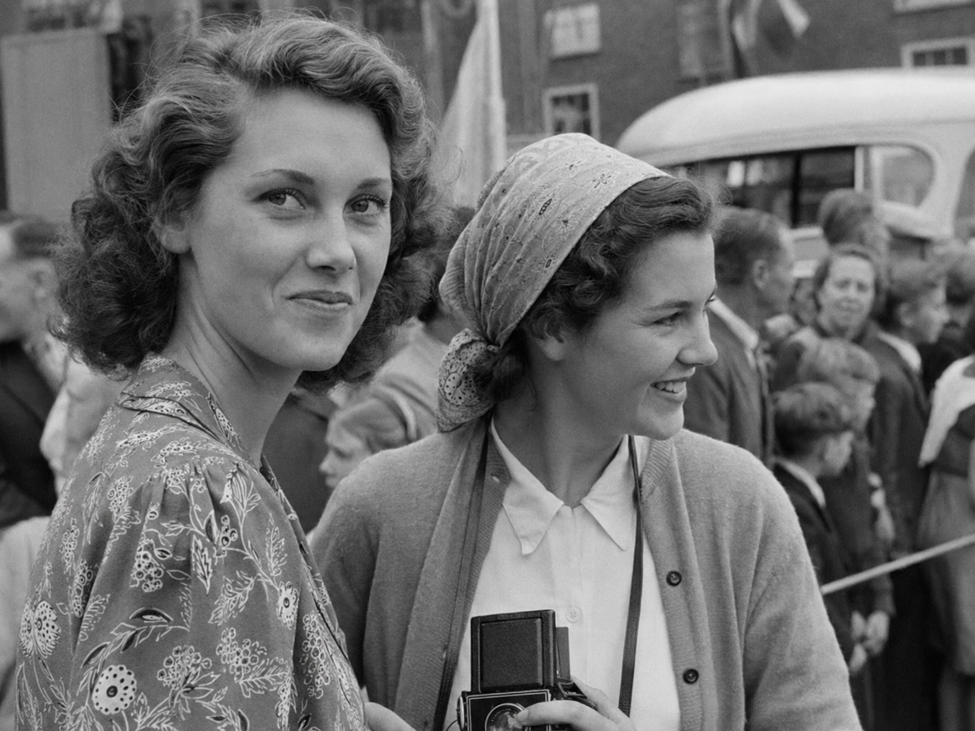 Two young women in the Nijmegen welcome crowd in June 1949, one looking toward the photographer while the other smiles toward the procession and holds a box camera.