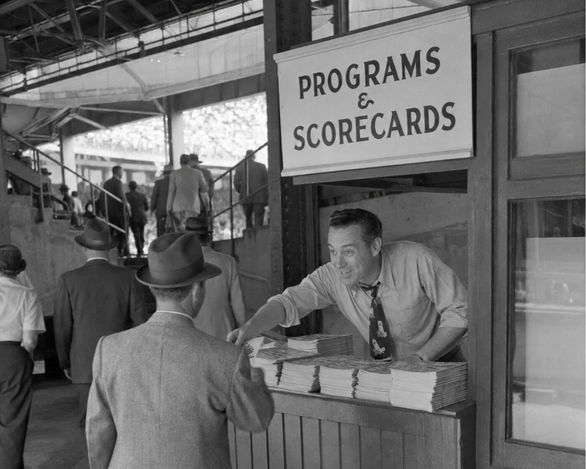 1940s ballpark vendor reaches across a counter stacked with scorecards toward a spectator beneath a Programs & Scorecards sign.
