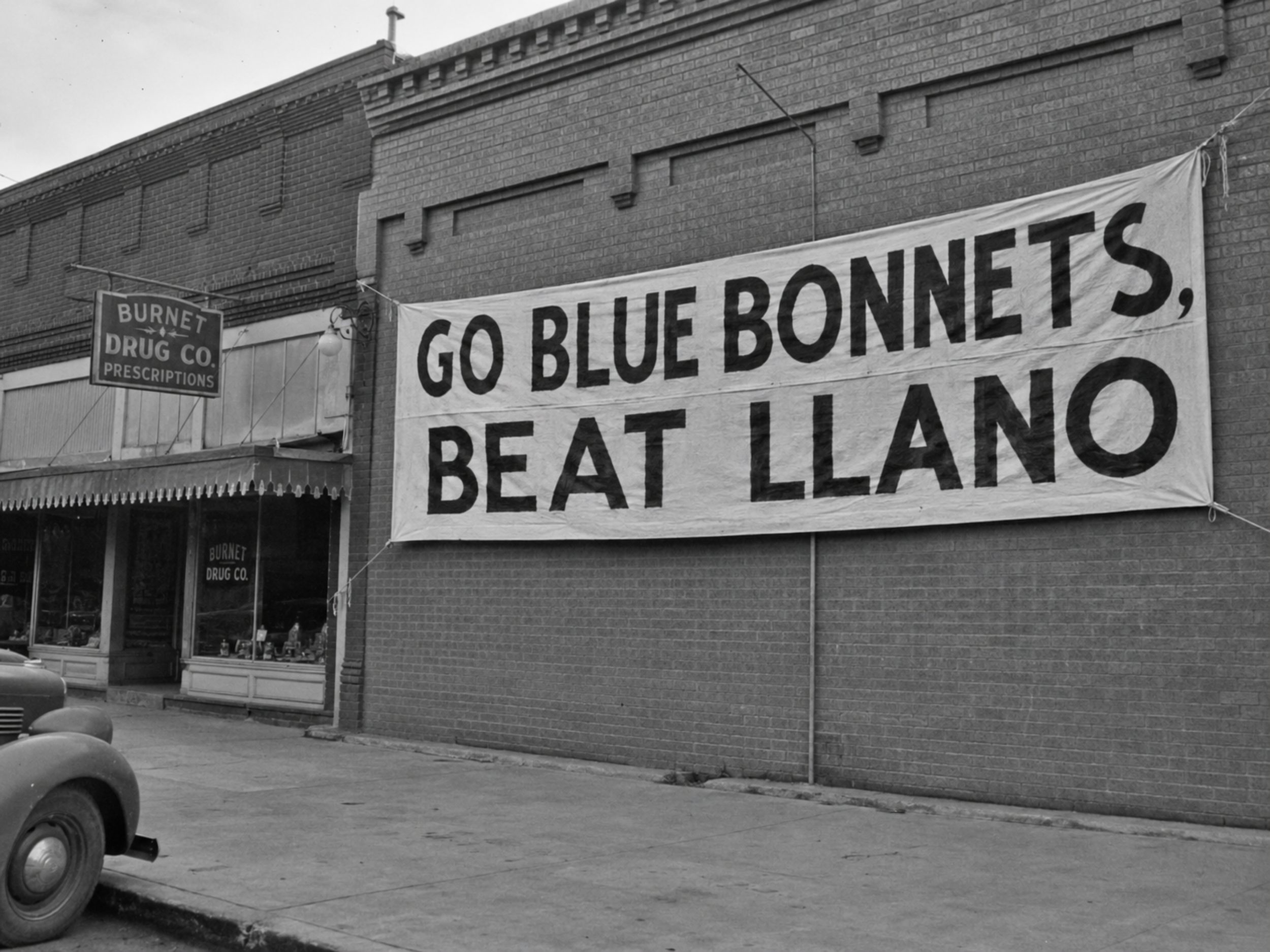 Black-and-white 1940s photograph of a large banner reading “GO BLUE BONNETS, BEAT LLANO” hanging on a brick building in downtown Burnet.