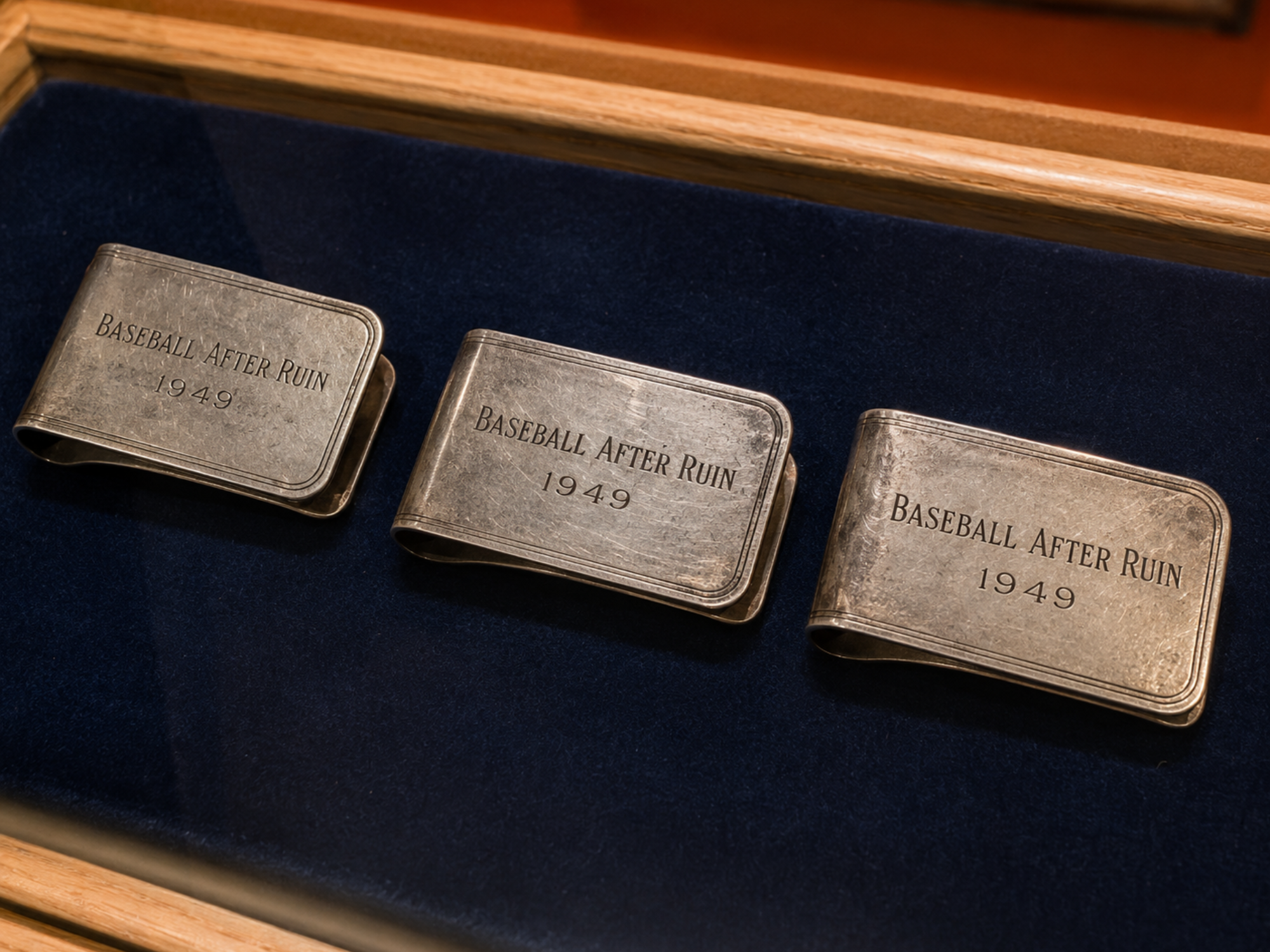 Three worn silver money clips engraved “Baseball After Ruin” and “1949” rest on Dutch navy velvet inside a blonde-wood museum case.