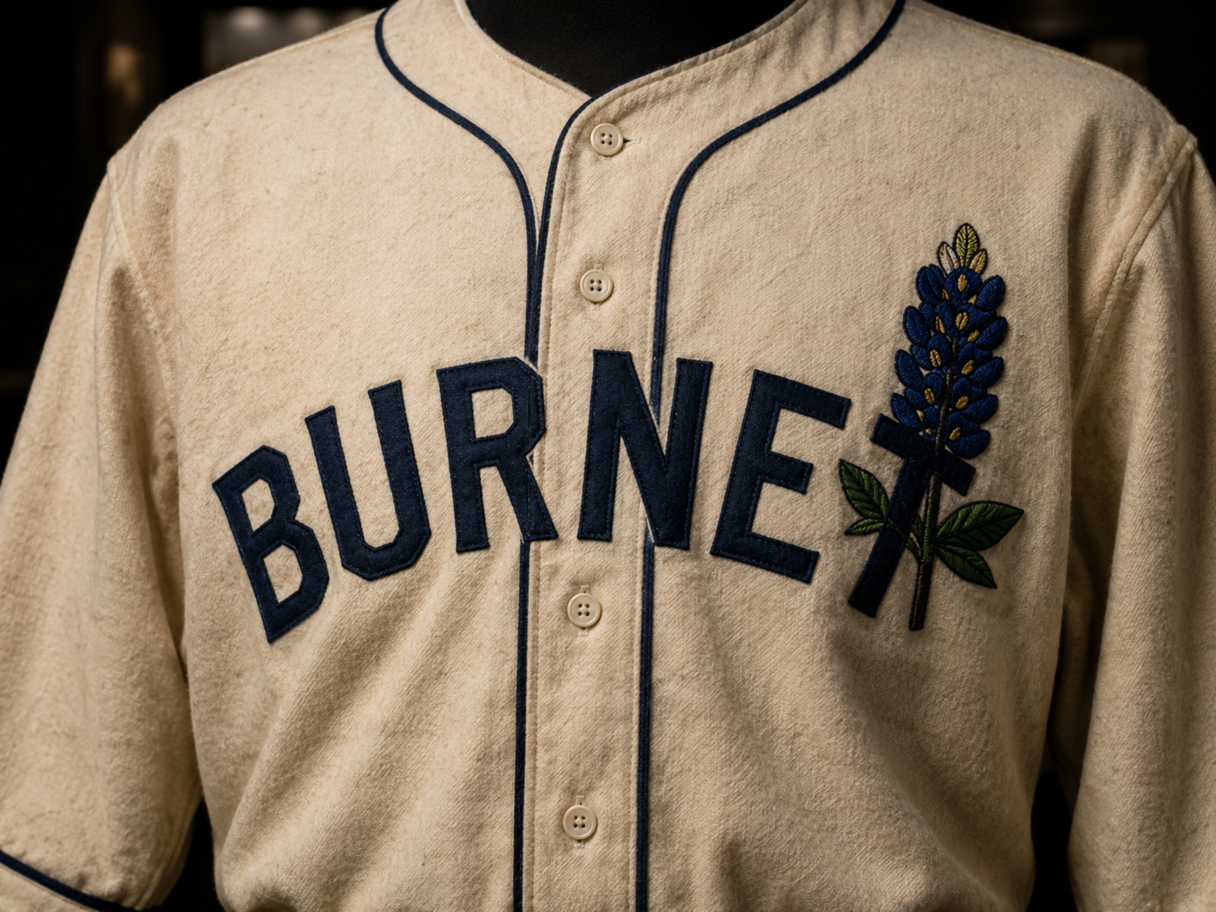 Close-up of a cream Burnet Blue Bonnets primary jersey showing dark-blue BURNET lettering, dark-blue piping, buttons, and a stitched bluebonnet chest emblem.