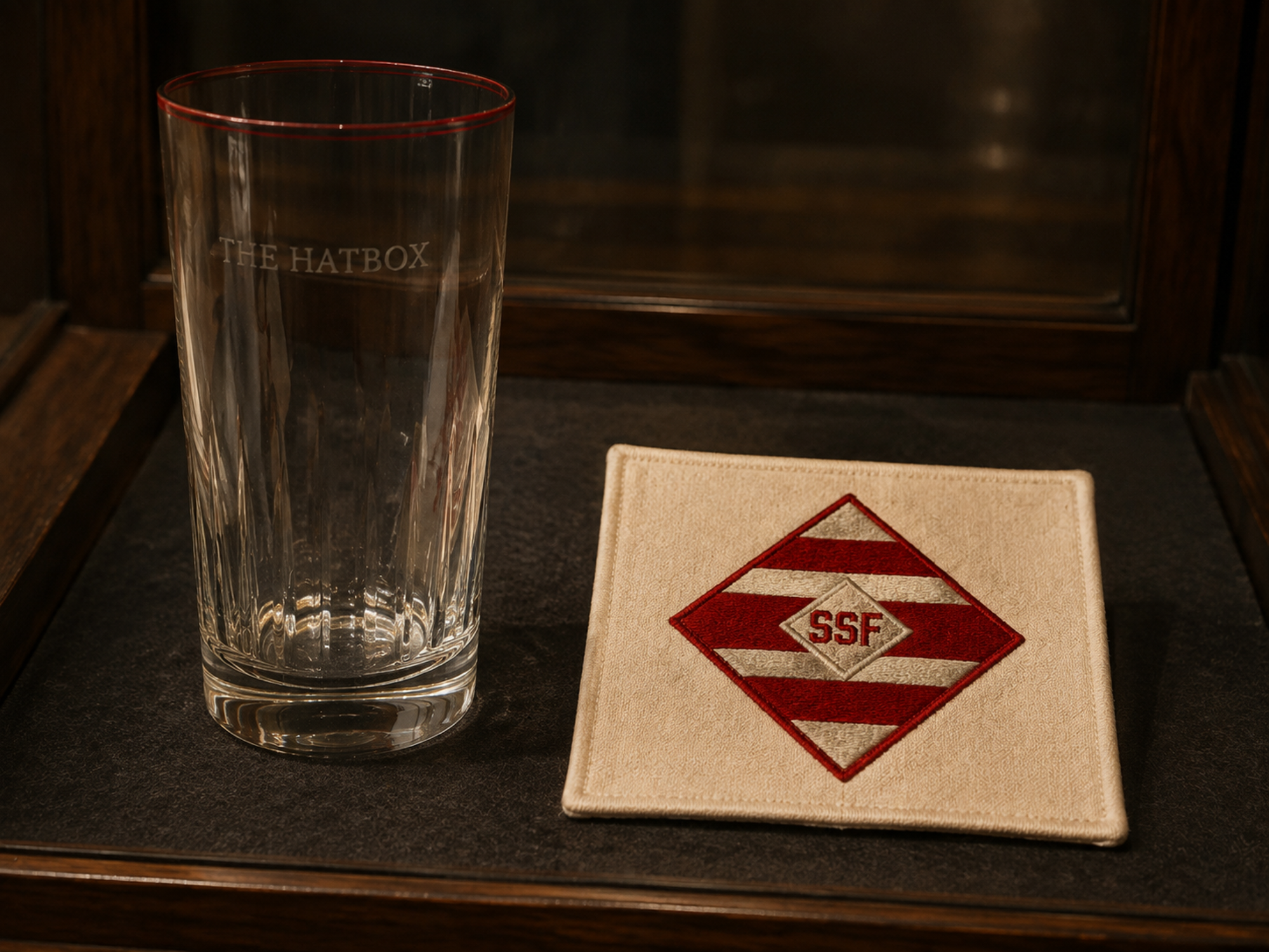 A preserved Hatbox Cup glass and embroidered SSF linen coaster displayed together in the museum.