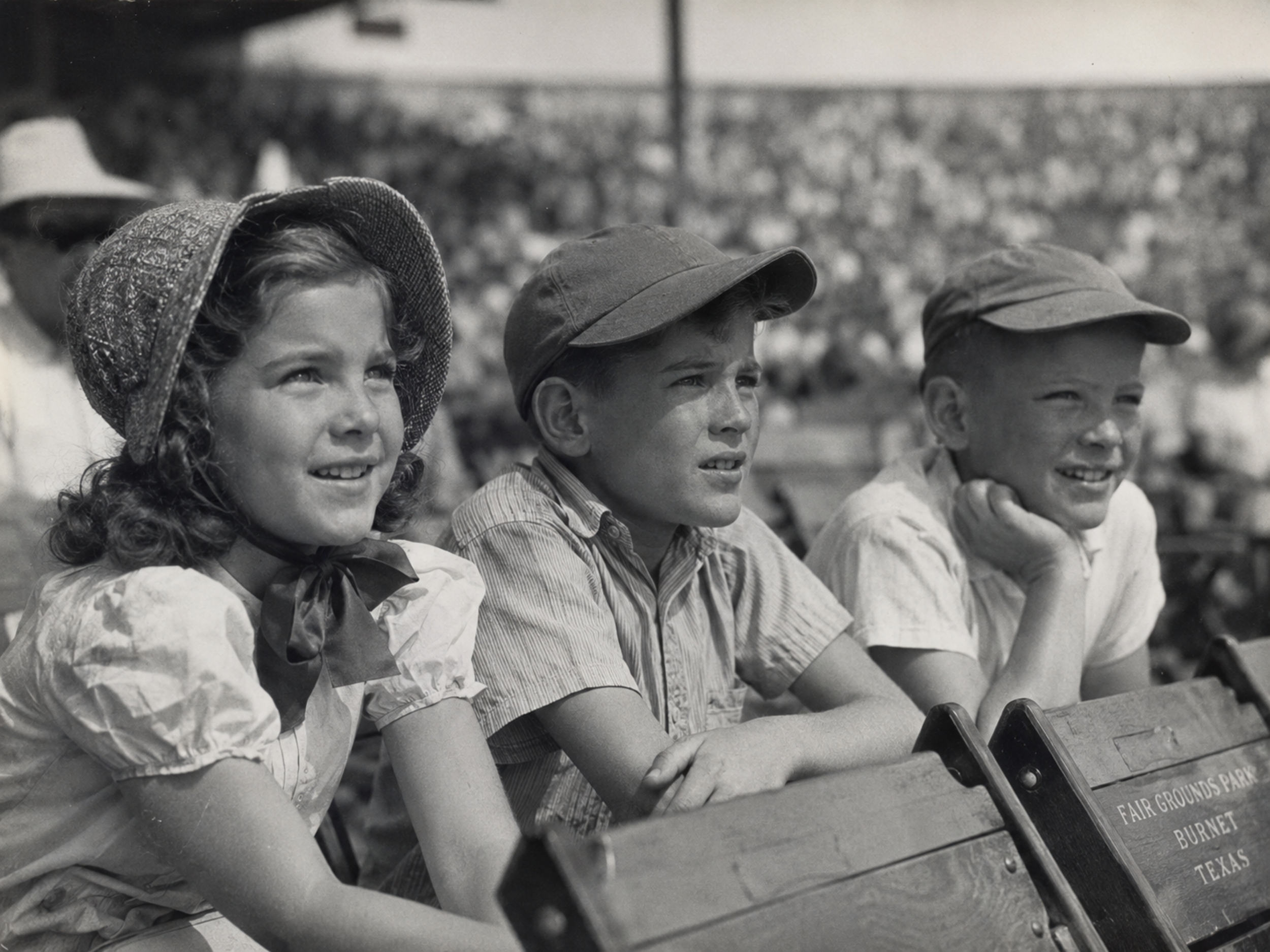 Black-and-white 1940s photograph of a young girl in a bonnet-style hat seated with two boys at Fair Grounds Park, watching a Burnet Blue Bonnets game with the crowd blurred behind them.
