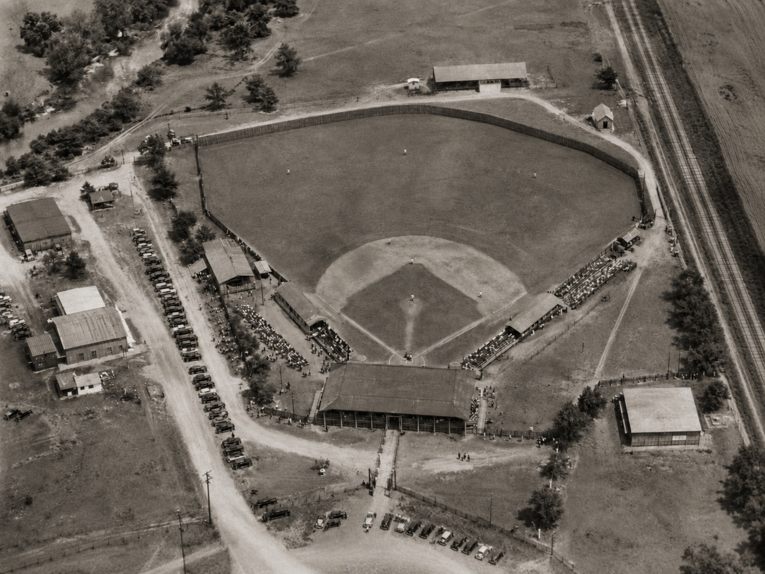Black-and-white aerial view of Fair Grounds Park in Burnet, Texas, showing the baseball diamond, roofed grandstand, first-base bleachers, outfield fence, nearby roads, parked cars, and railroad siding.