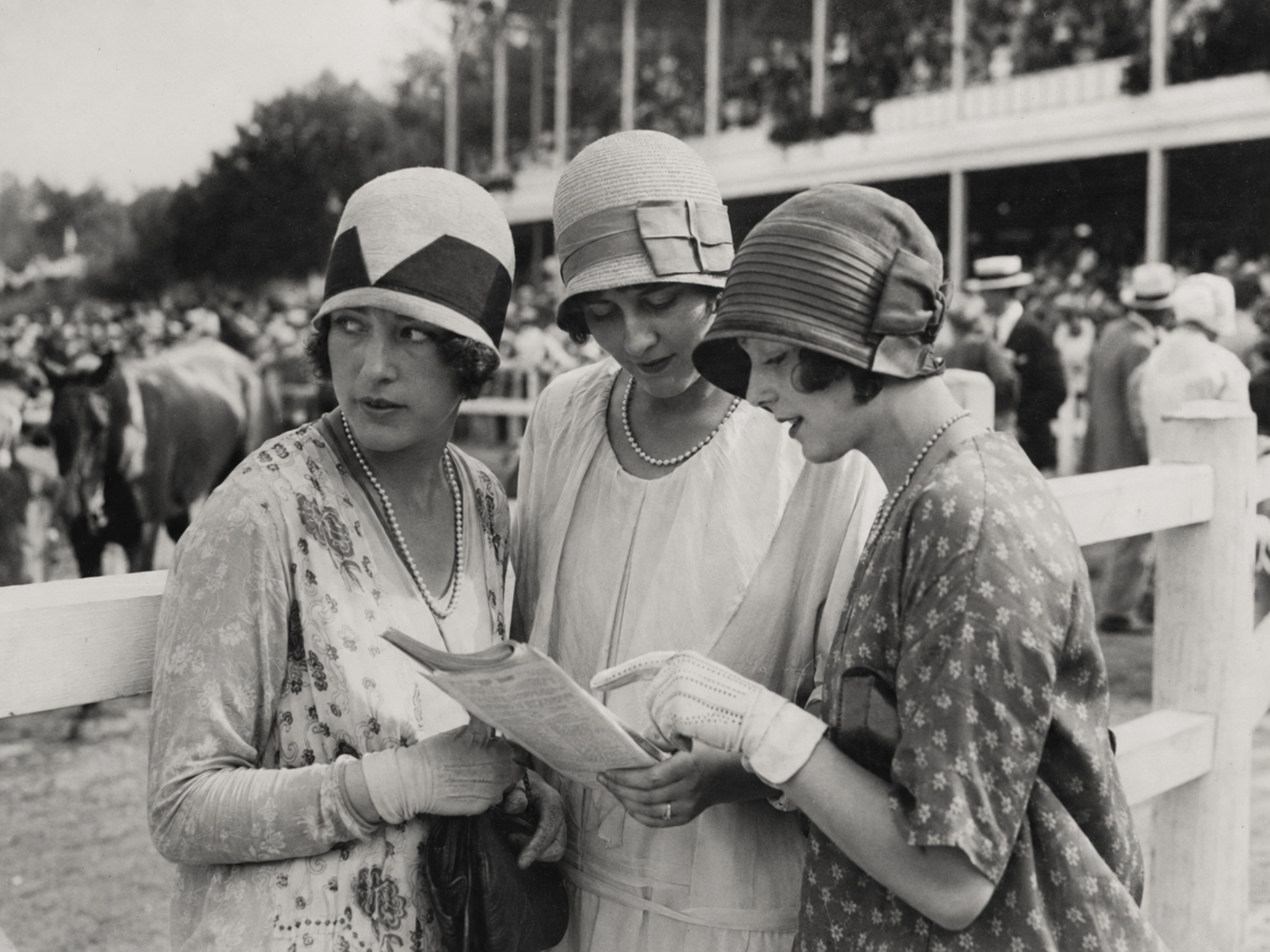 Three women in clothe hats studying a race program near the Saratoga paddock in 1927.