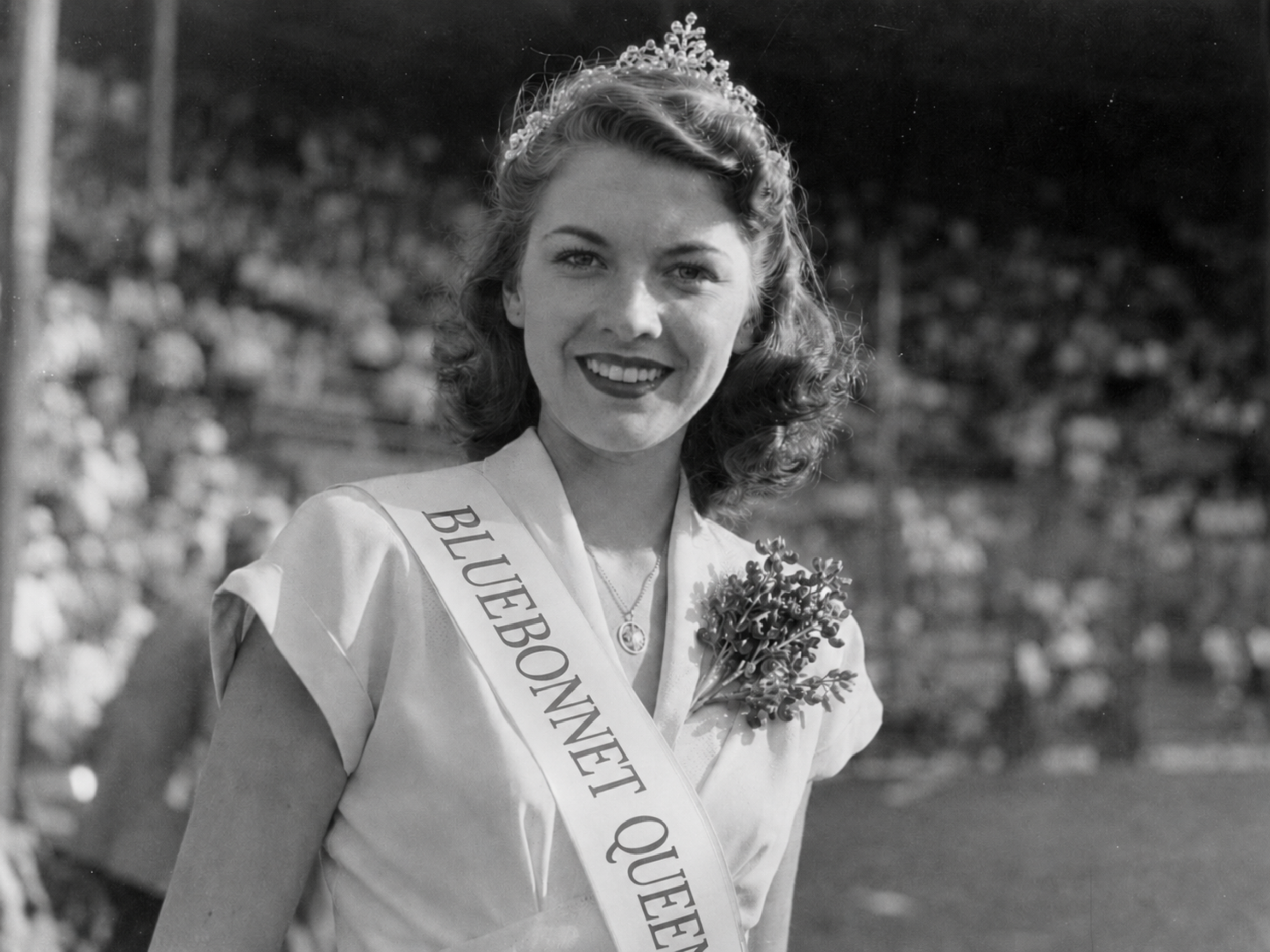 Black-and-white 1940s portrait of the Bluebonnet Queen smiling at a Burnet Blue Bonnets game, wearing a sash and floral corsage with the Fair Grounds Park crowd blurred behind her.