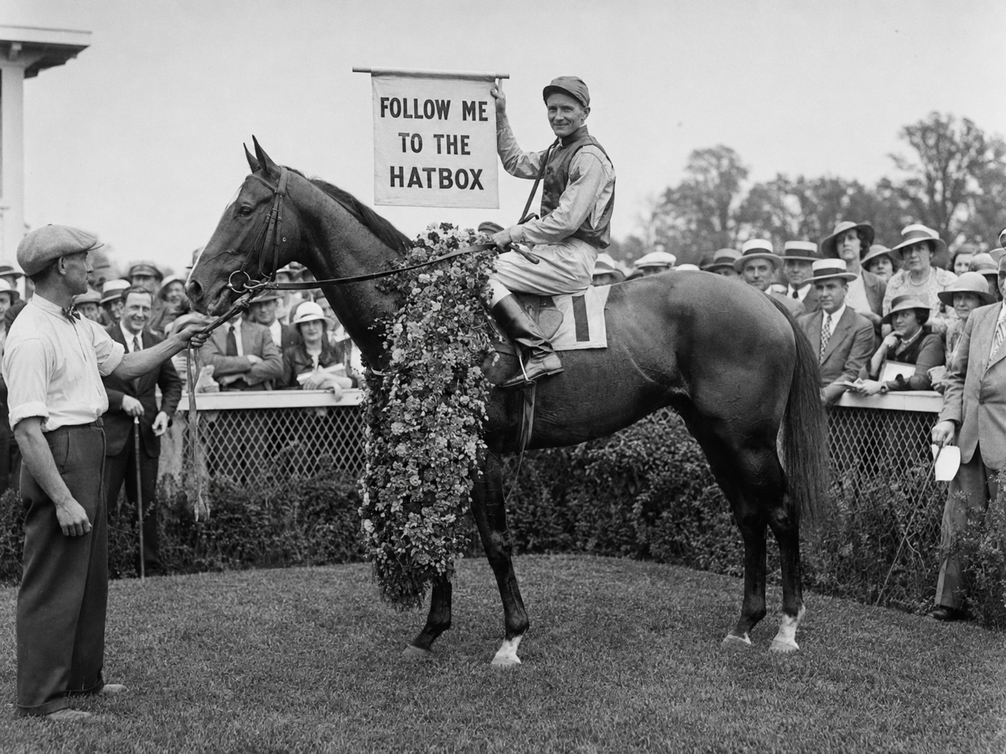A jockey aboard a winning horse holds a “Follow Me to The Hatbox” banner in the winner’s circle at Saratoga Race Course.