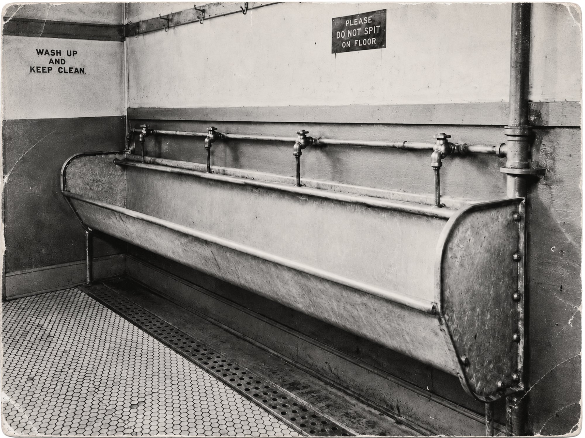 Black-and-white 1940s photograph of a metal trough urinal with exposed plumbing, hexagonal tile, coat hooks, and hand-painted signs beneath the West End Park grandstand.