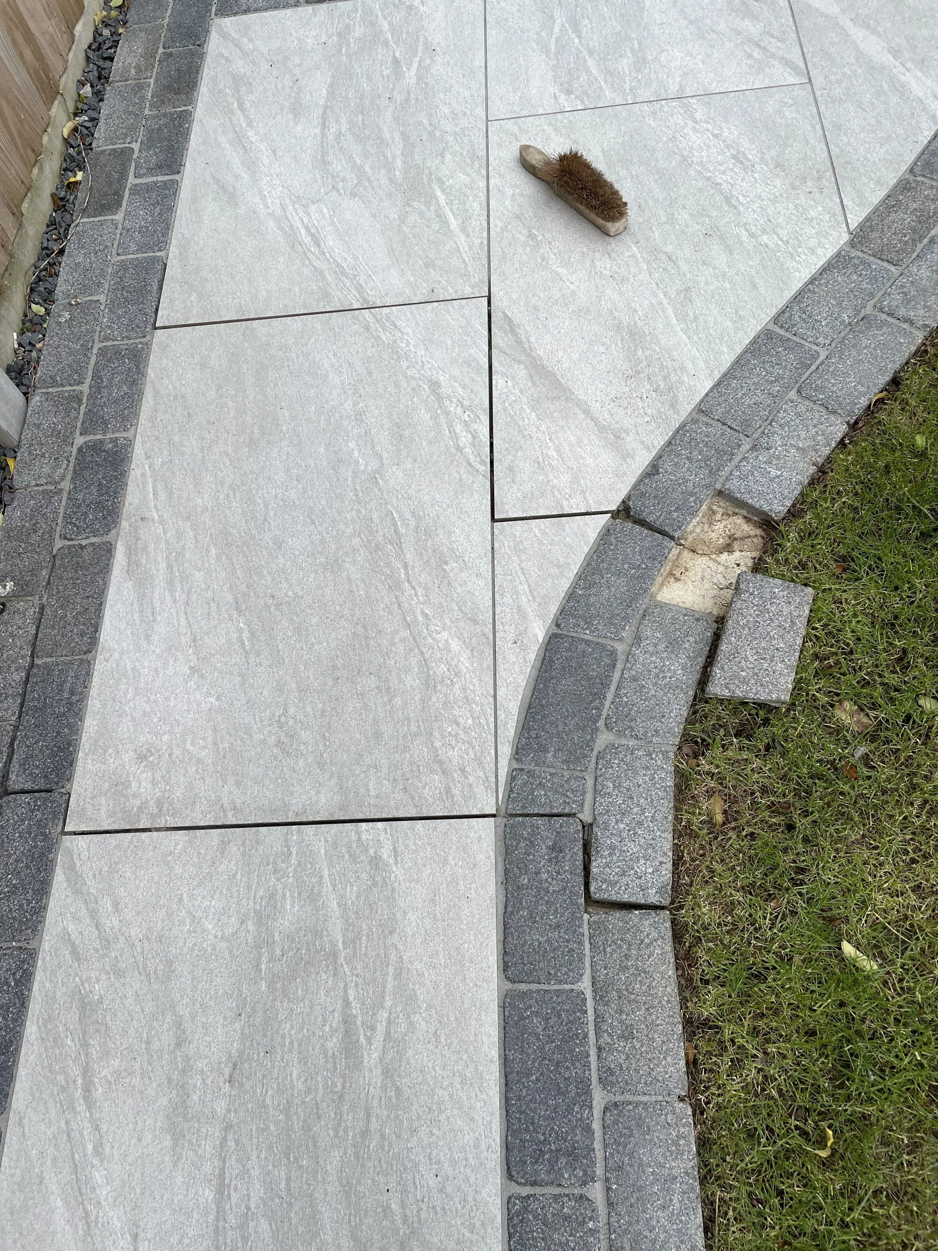 Close-up of a section of a tiled outdoor walkway bordered by a row of gray bricks and a grassy area with a small flower.