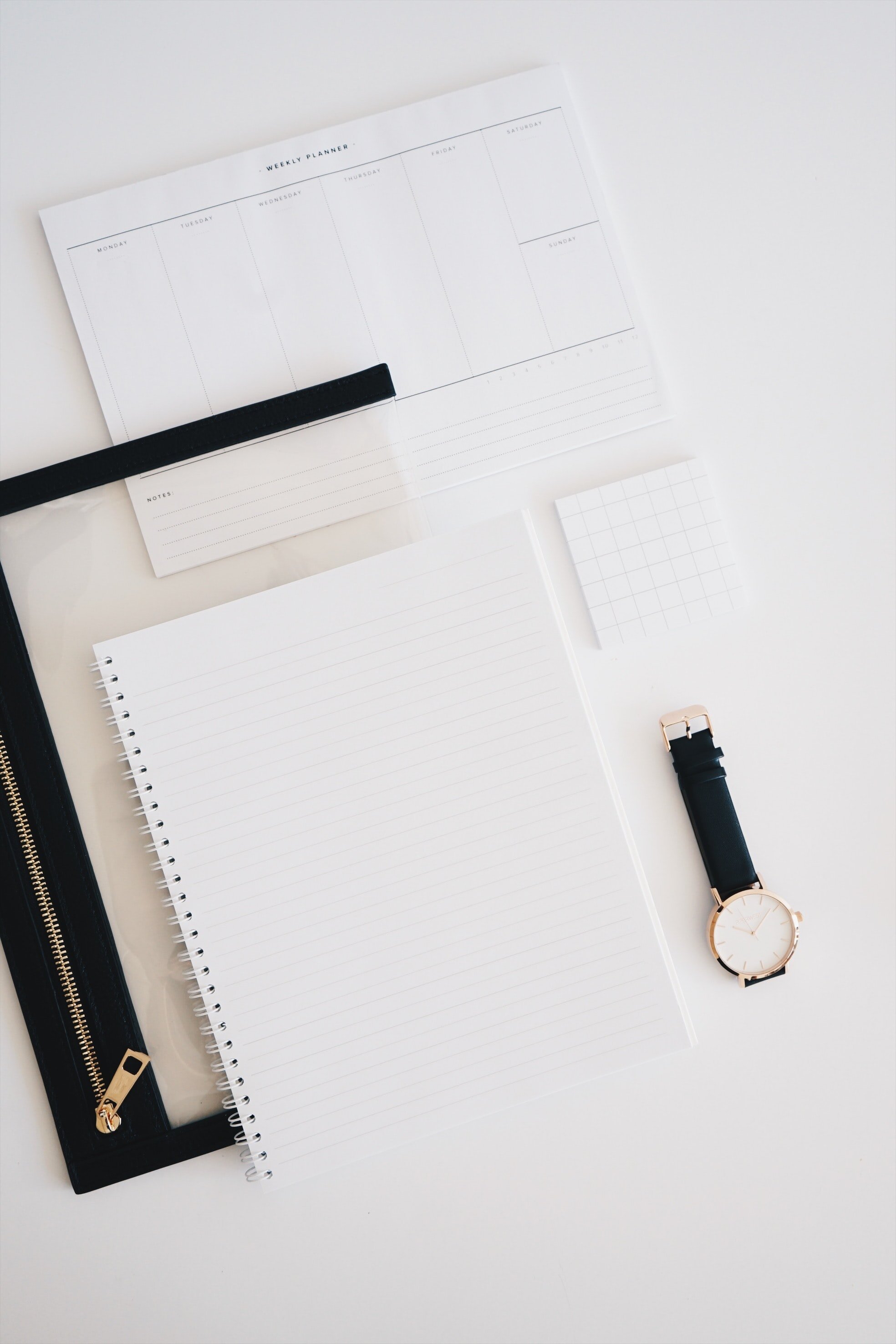 A flat lay of an open weekly planner with no entries, a black pen, a small notepad, a wristwatch with a black strap and gold accents, on a white desk surface.
