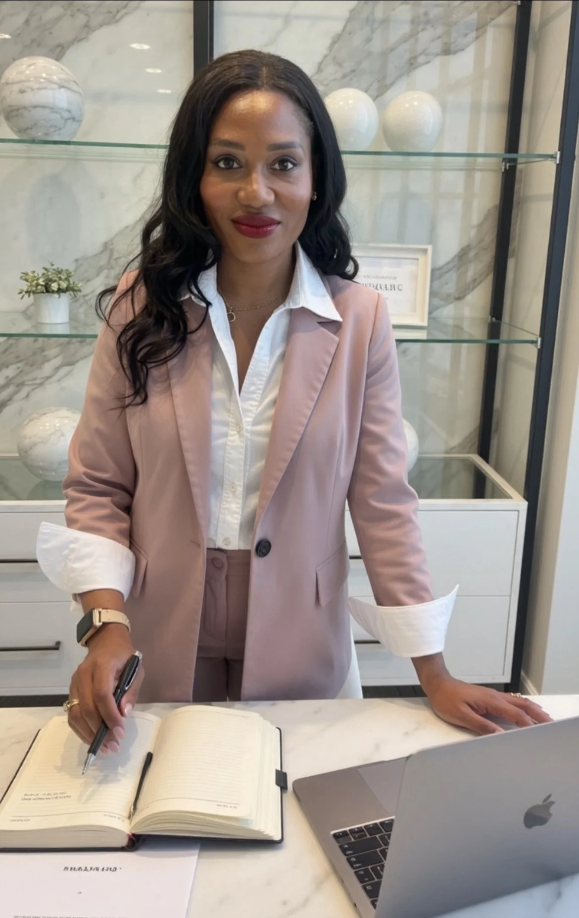 A woman with long dark wavy hair, wearing a pink blazer over a white button-up shirt, and red lipstick, standing in front of a marble shelf with white decorative spheres. She is holding a pen over an open notebook on a white marble table and a laptop nearby.