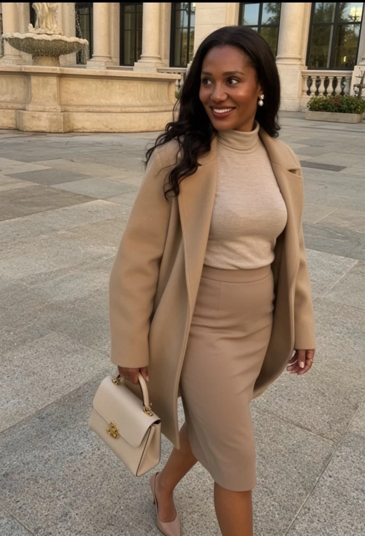 A woman dressed in beige clothing standing outdoors near a fountain and historic building, carrying a beige purse.