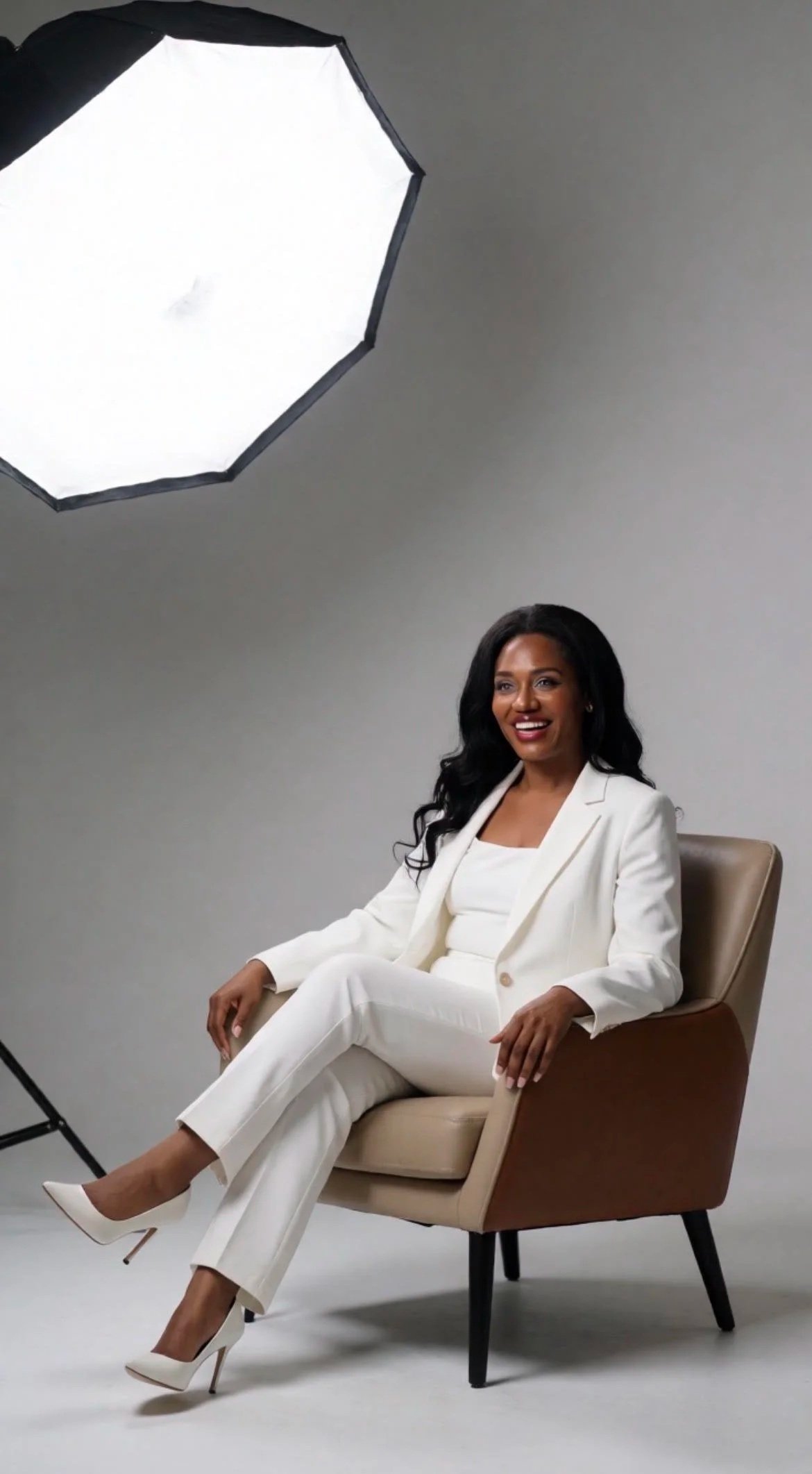 Sanjanetta Barnes in a white suit sitting on a beige armchair during a photoshoot, with professional studio lighting equipment overhead.