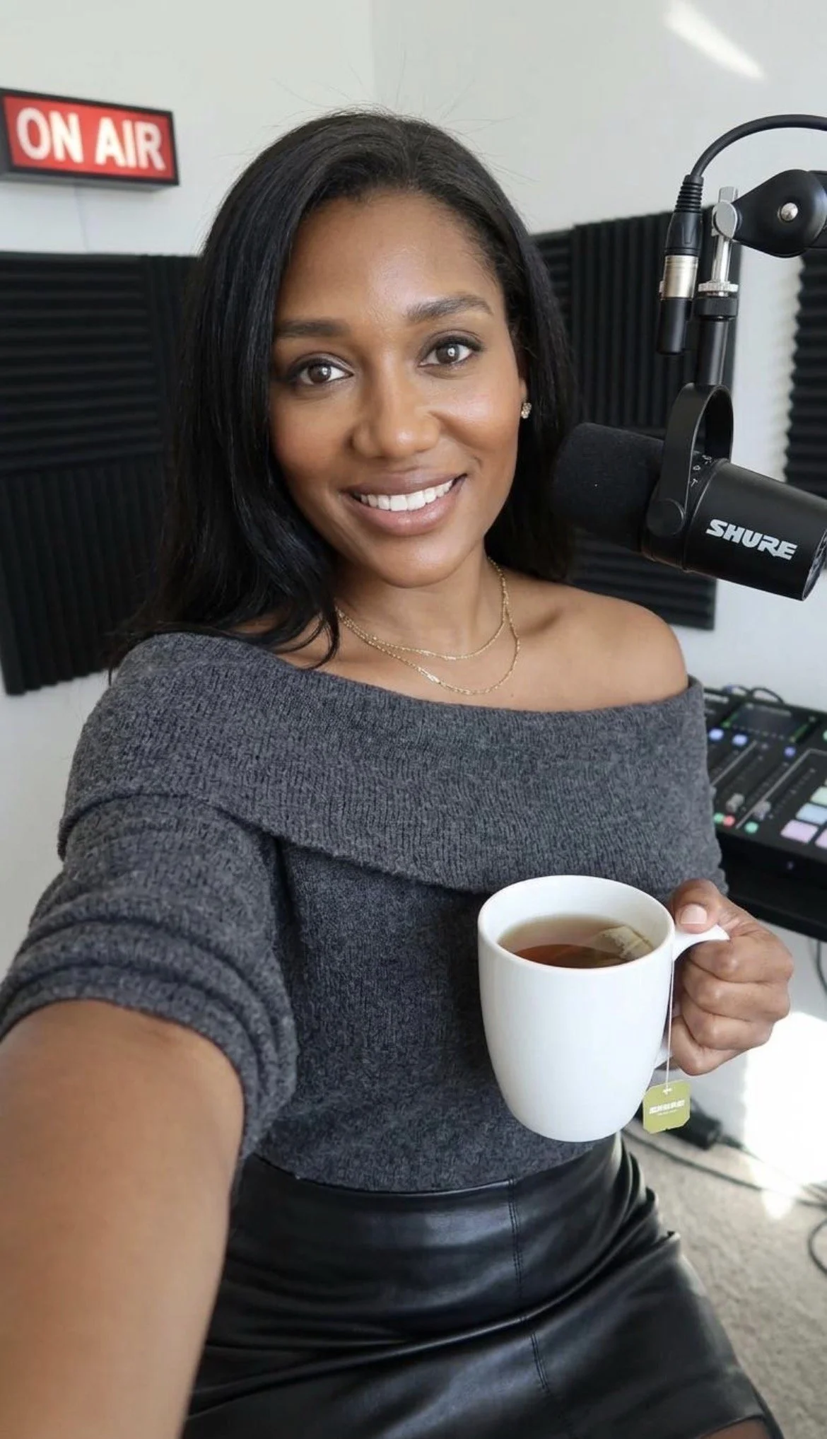 A woman in a studio with radio equipment and soundproofing, holding a mug of tea, smiling at the camera.