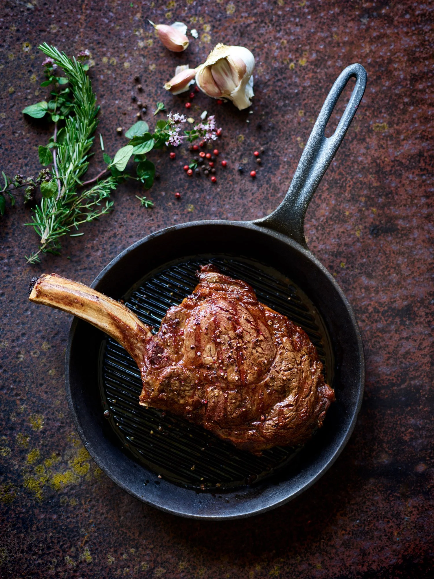 griddled tomahawk steak on the bone with garlic, rosemary bay and peppercorns on a dark rich background