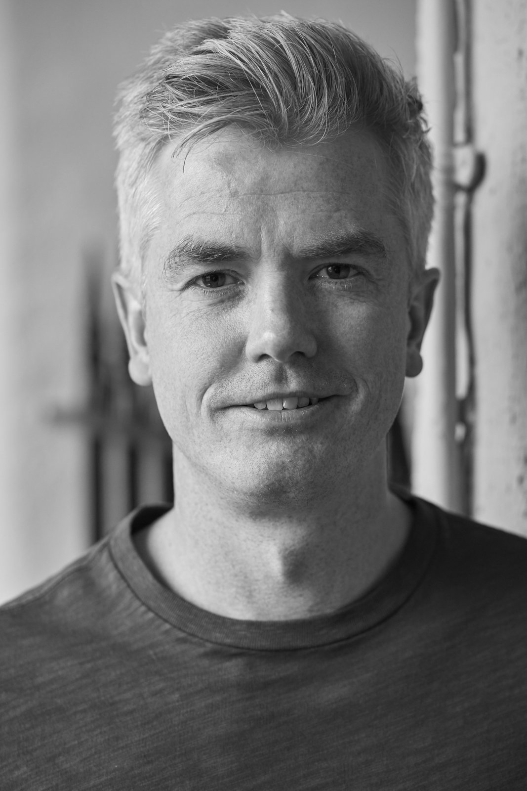 Black and white close-up photo of a smiling young man with short, styled hair, wearing a t-shirt, standing against a blurred background.