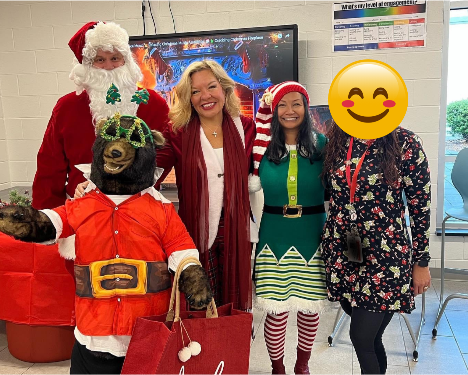 Group of five people dressed in Christmas costumes and holiday attire, smiling and posing for a photo indoors with Christmas decorations and a holiday-themed background behind them.